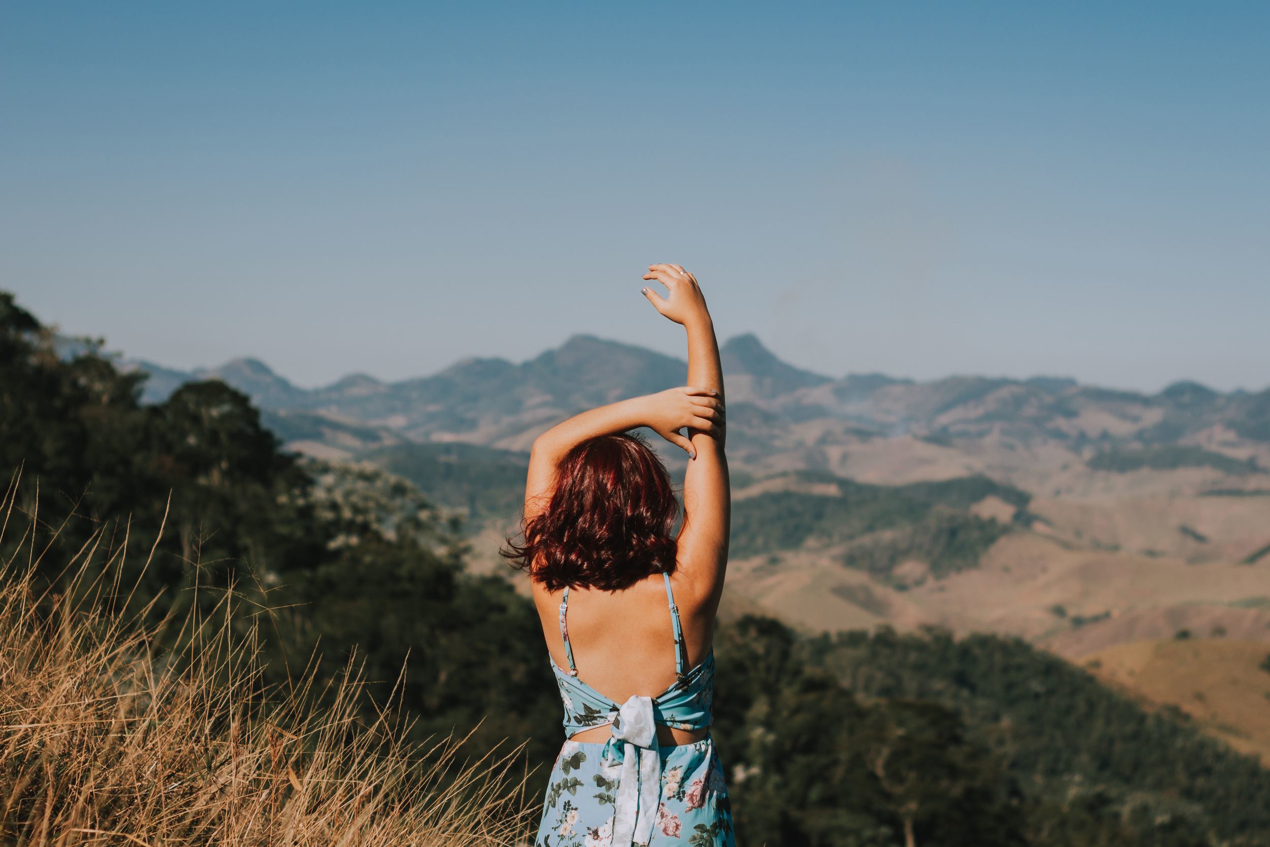 A woman standing with her right arm raised being supported by her left arm which is resting on her head overlooking a mountain range