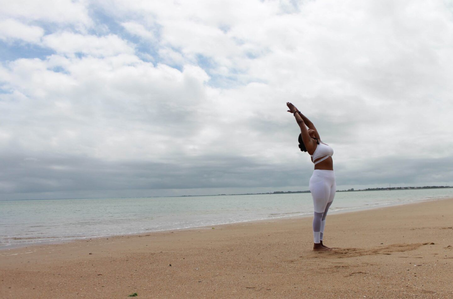 A woman is standing on an empty, peaceful beach. She's wearing white exercise clothes and stretching, greeting the day.