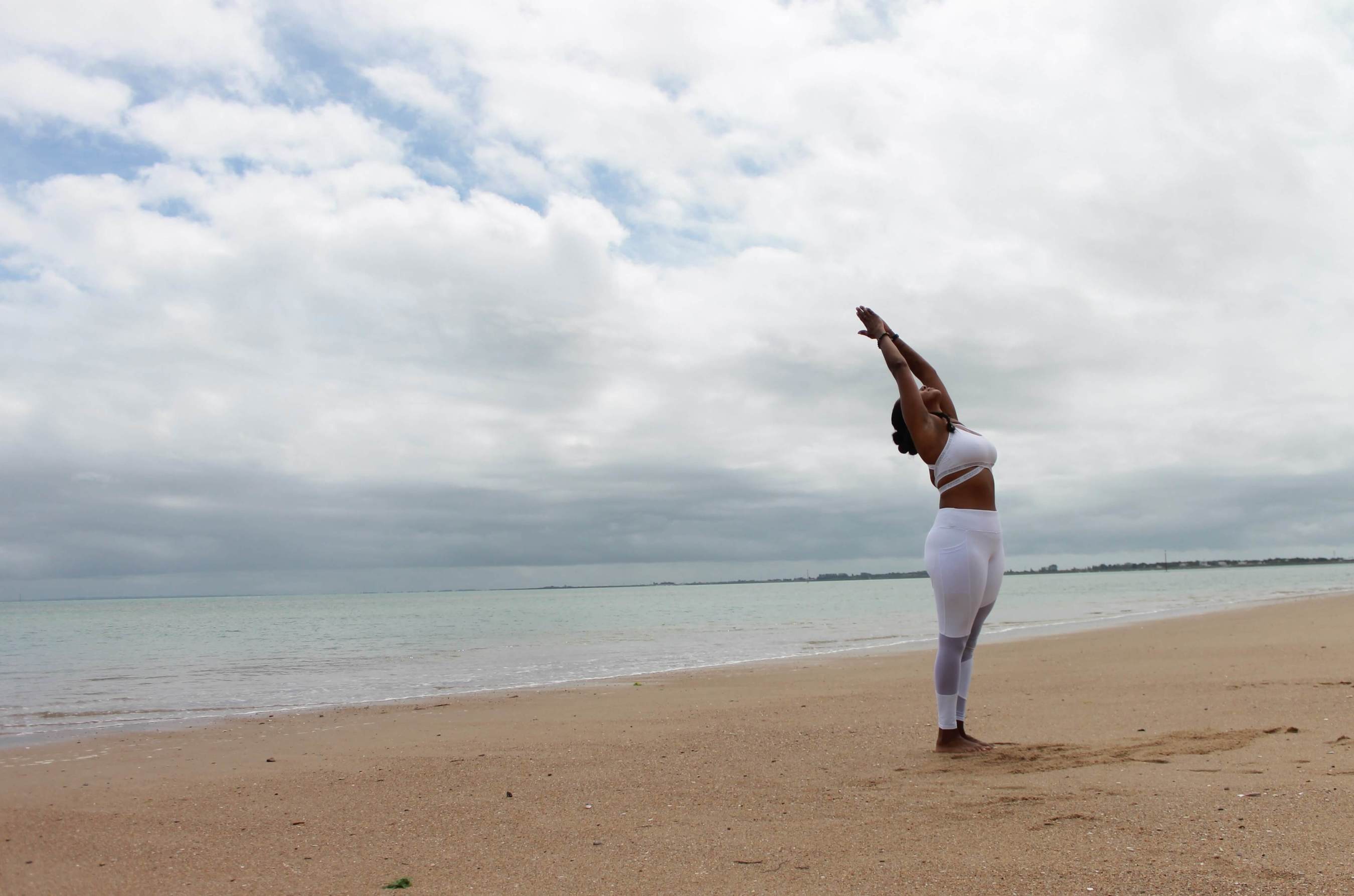 A woman is standing on an empty, peaceful beach. She's wearing white exercise clothes and stretching, greeting the day.