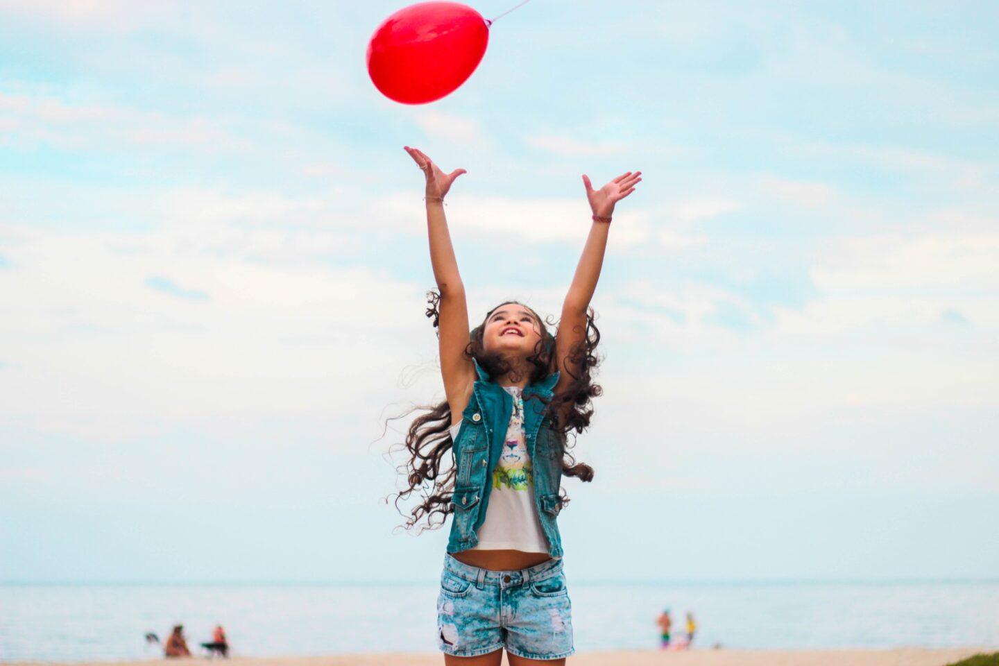 A young kid enjoying their day on the beach, chasing after a red balloon