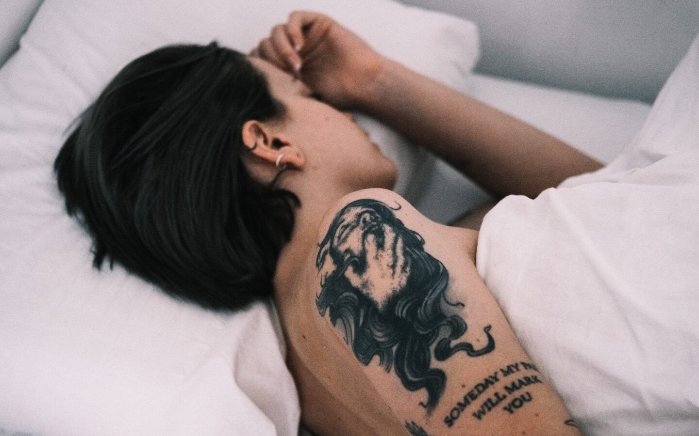 Tattooed person with short brown hair is sleeping in a cozy bed with white sheets