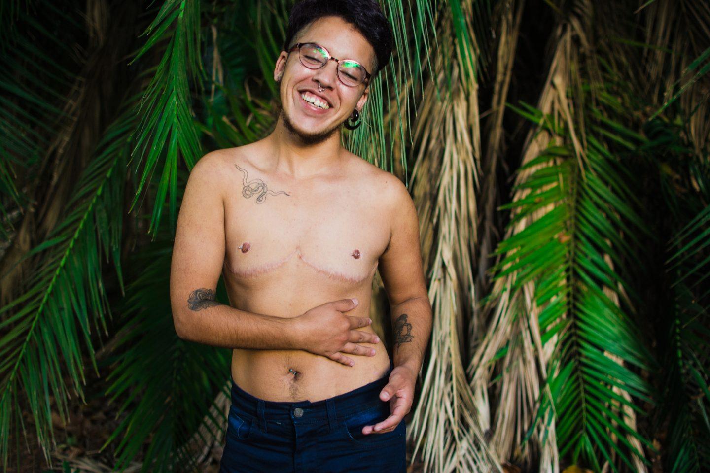 Young transgender man portrait in a nature environment wth tree leafs green and dried. He is standing among the leaves wearing jeans and bare chest.