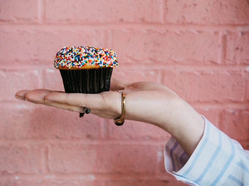 A hand holding a pink frosted cupcake with sprinkles against a pink wall