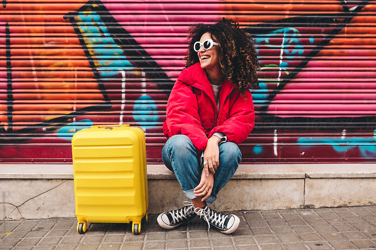 a black woman smiling sitting on a curb with her yellow suitcase