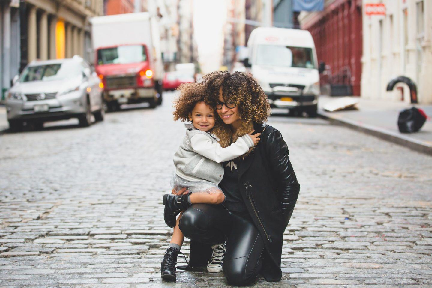 a black woman with her child kneeling in a cobble alley with parked cars behind them