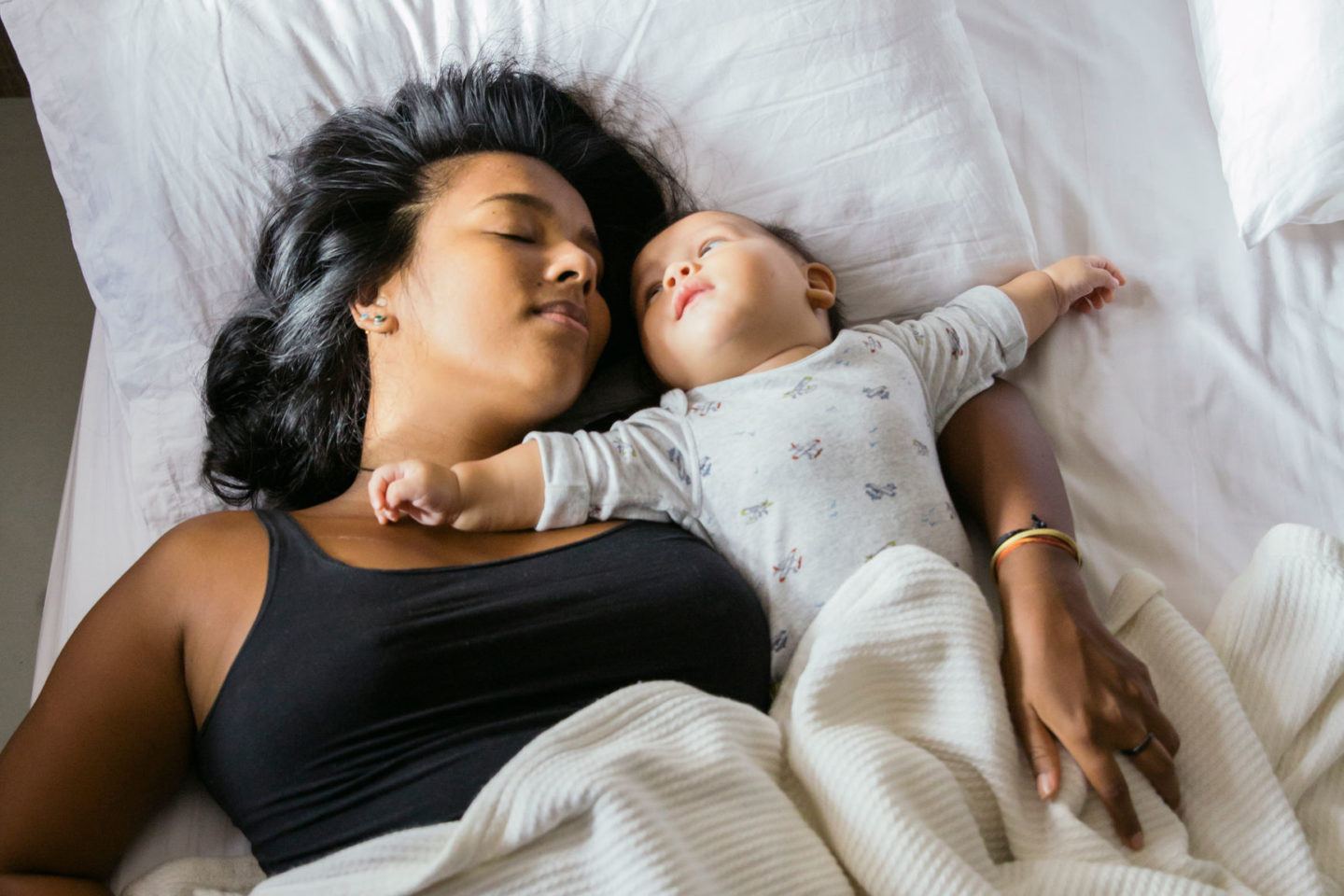 A mother and her baby lie in bed together on crisp white sheets in the early morning