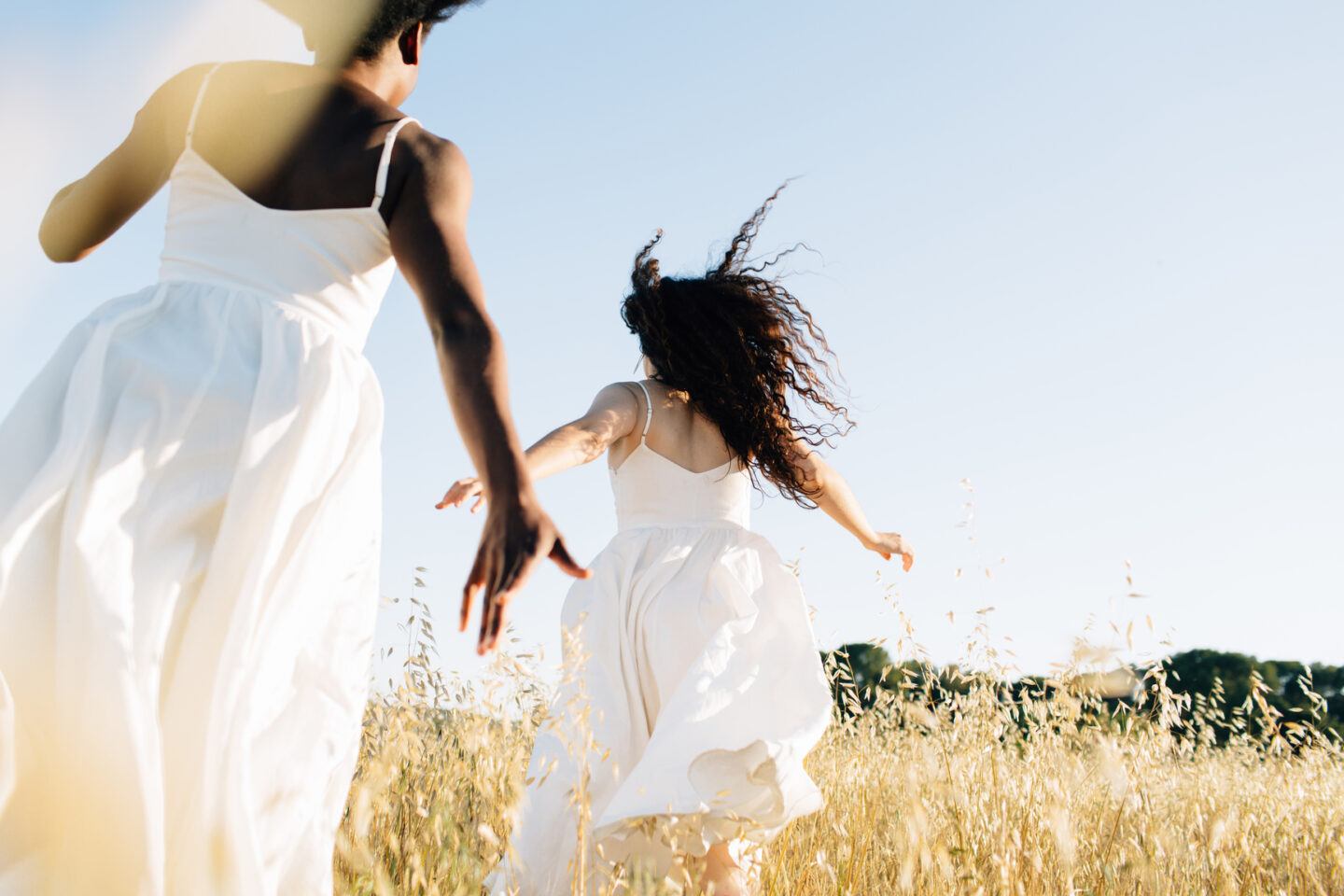 Two girls from behind , running in the middle of a dry field they wearing maxi dresses.