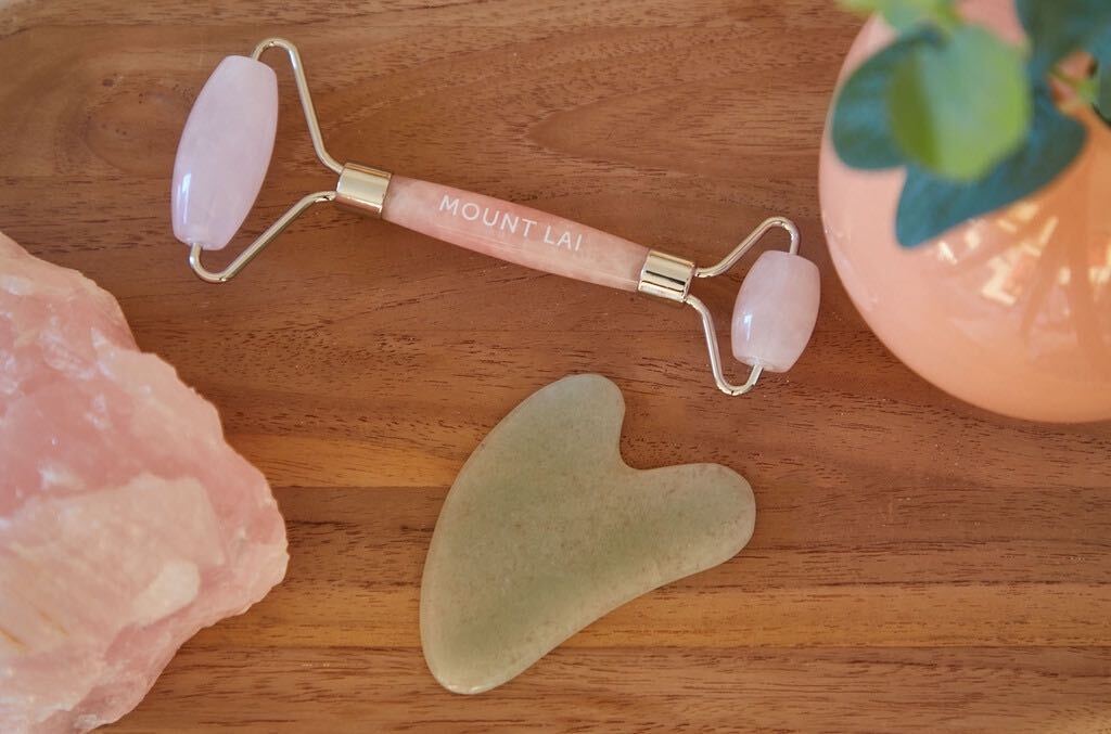 A rose quartz facial roller and a jade gua sha tool lay on a wooden table between a rose quarts crystal and a peach vase