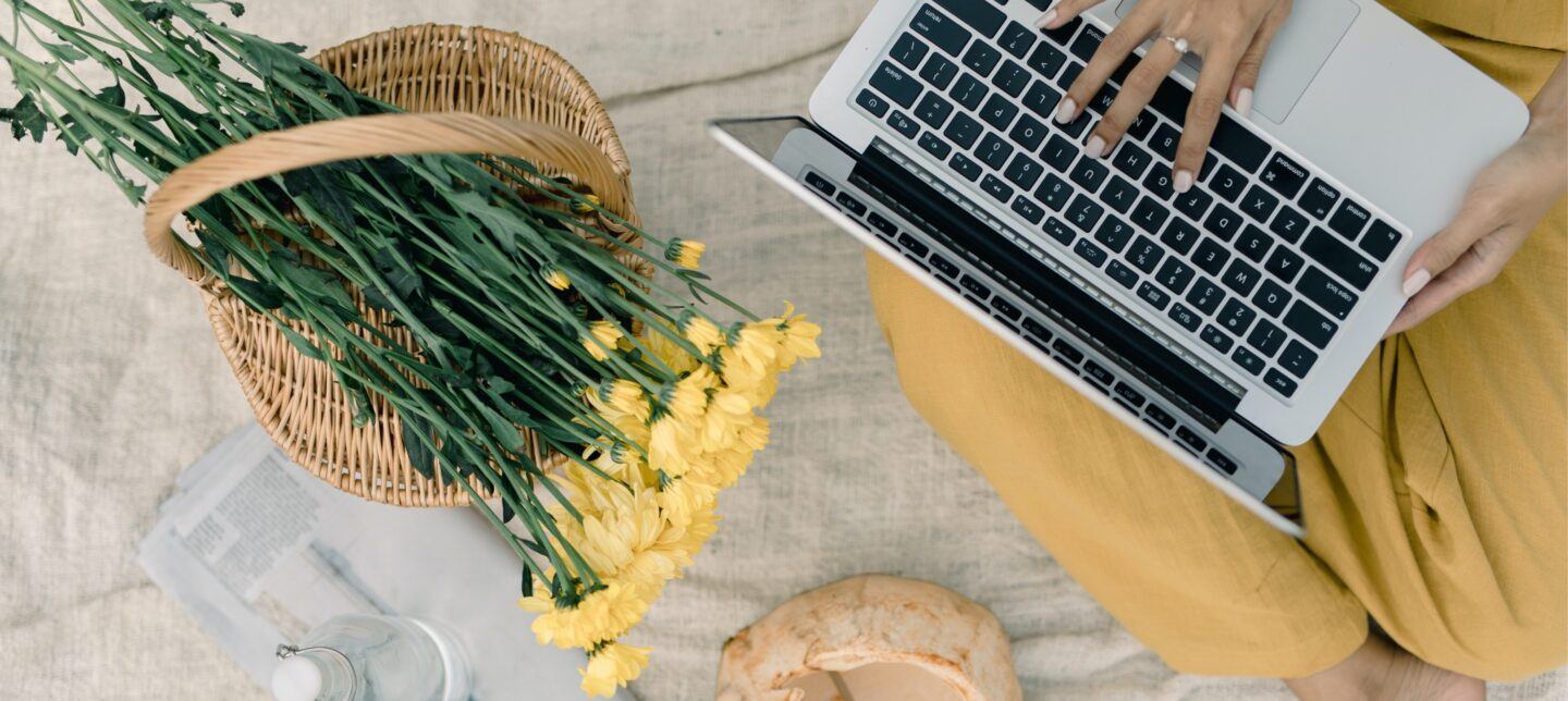 woman sitting on a blanket with her laptop next to a basket with flowers