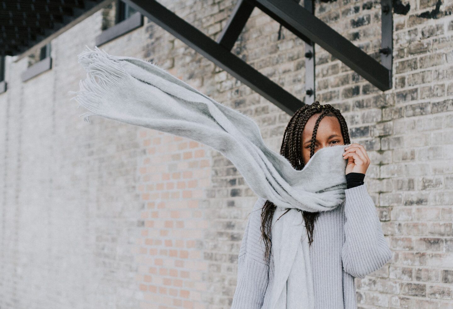 black woman with a gray scarf blowing in the wind standing against white brick wall