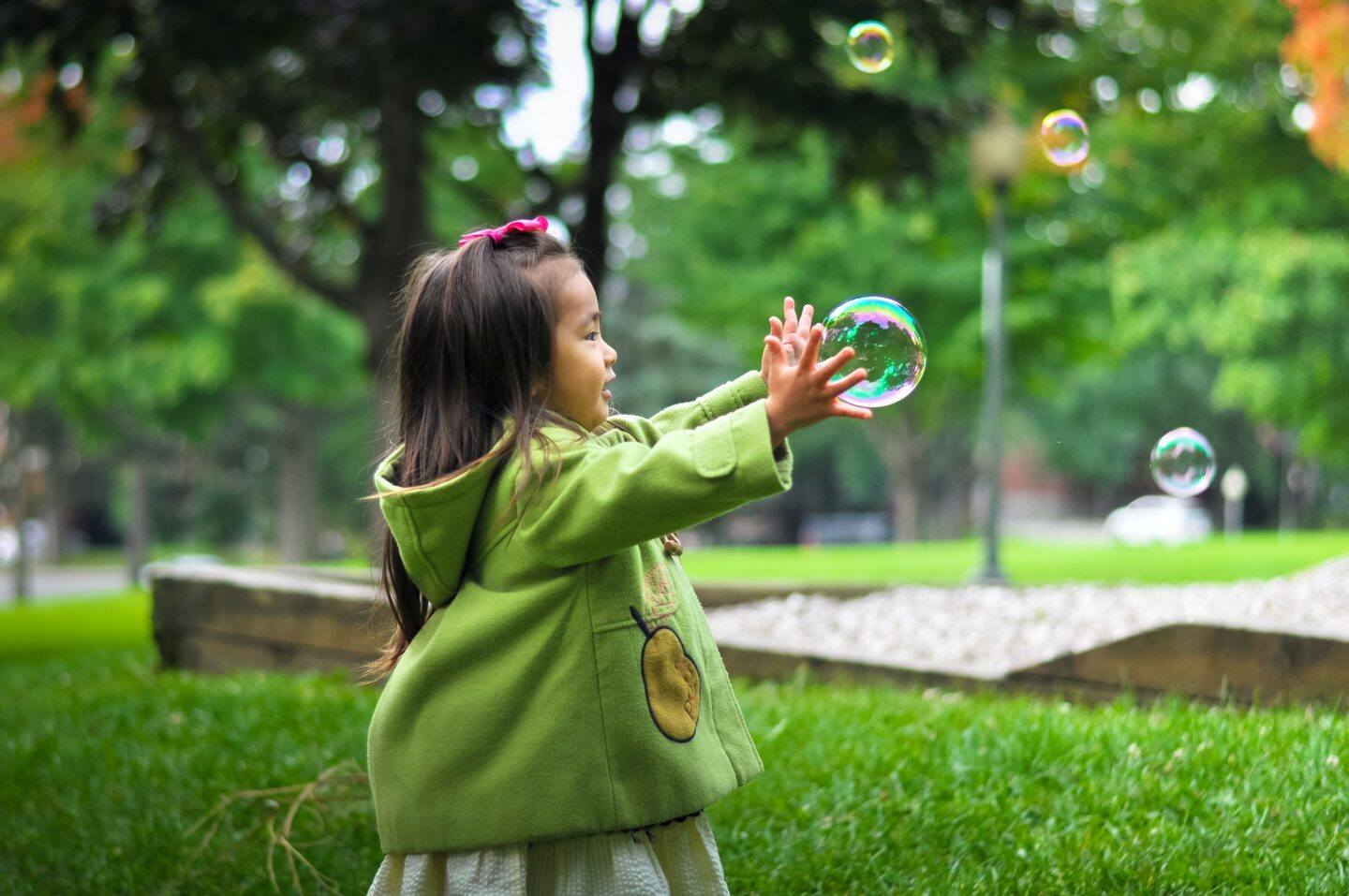 pictured is a little girl of Asian descent wearing a green coat playing with bubbles outside