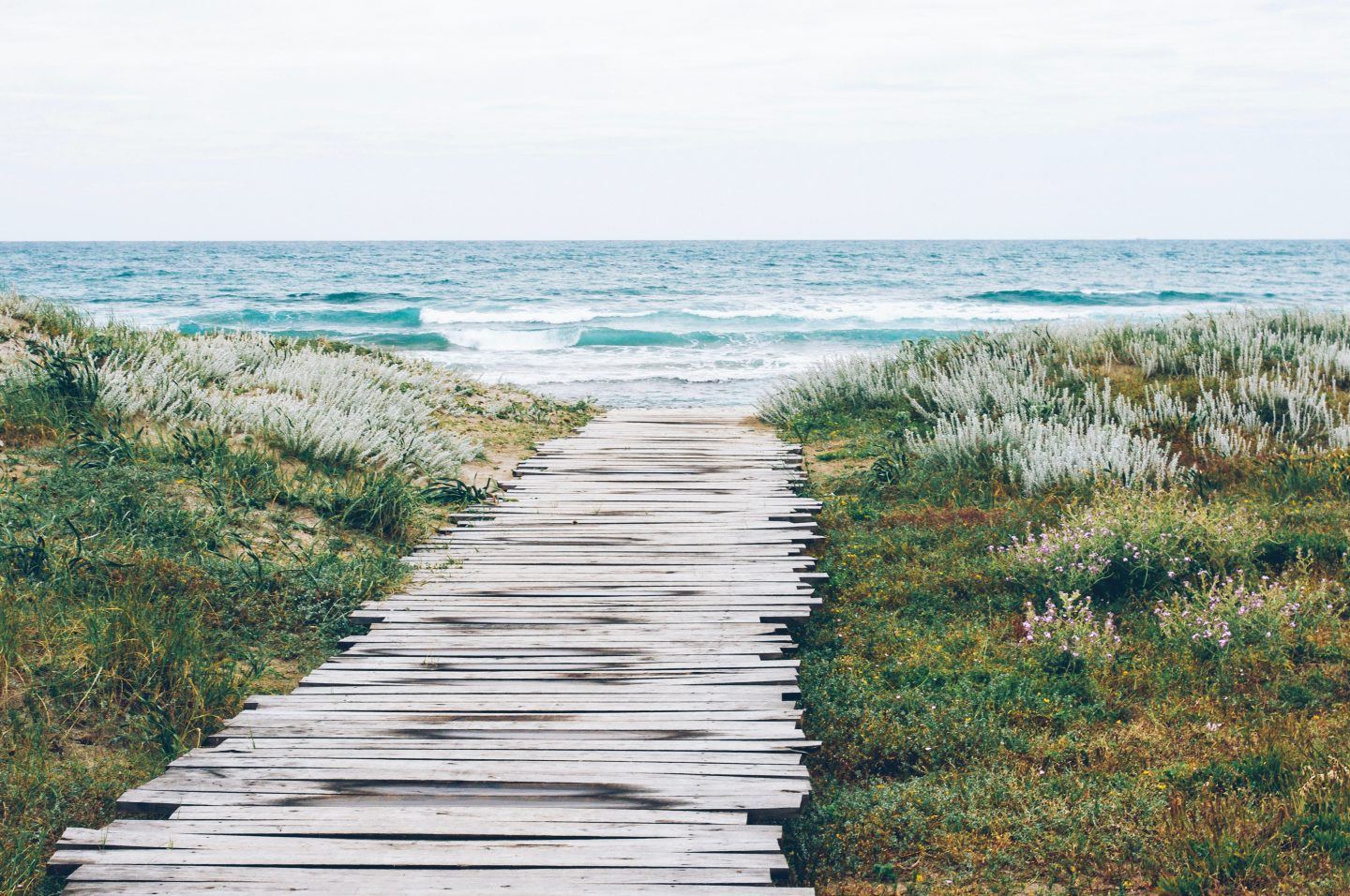 A wooden path surrounded by grass leads to the beach with crashing blue waves