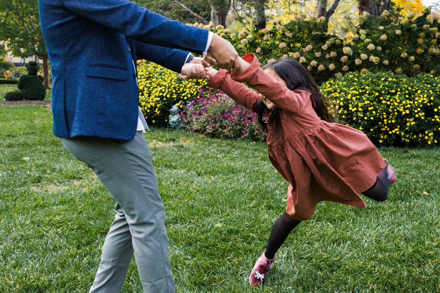 Parent dressed in blue swings their child around in a vibrant green garden, child is wearing a red dress