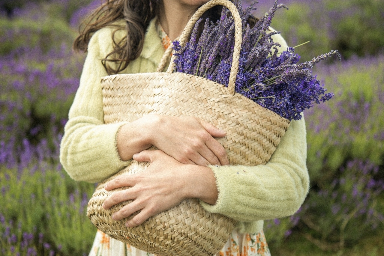 a woman in a green and purple lavender field holds a woven basket filled with lavender